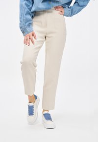 Pantaloni beige a vita alta con tasche laterali e dettagli pieghettati, abbinati a sneakers bianche con lacci blu e suola robusta.