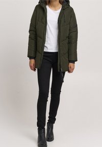 Pěnová olivově zelená parka s kapucí, zipovým zapínáním a postranními kapsami, v kombinaci s bílým tričkem a černými skinny džíny.