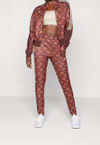 Brun idrottsdräkt med ett heltryckt palmönster, som har vita ränder och en dragkedja-jacka, tillsammans med vita sneakers.