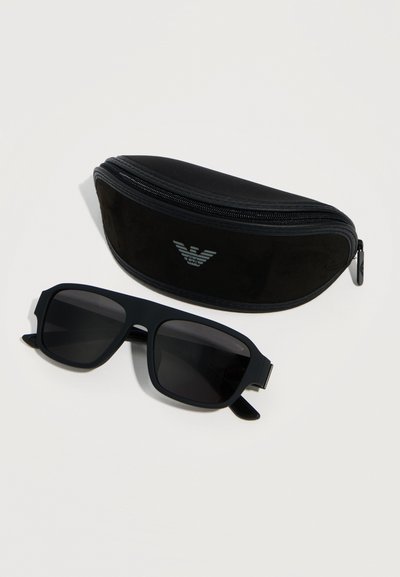 Μαύρα ορθογώνια γυαλιά ηλίου με σκούρους φακούς δίπλα σε μια μαύρη θήκη με φερμουάρ, που φέρει ασημένιο λογότυπο, σε λευκό φόντο.