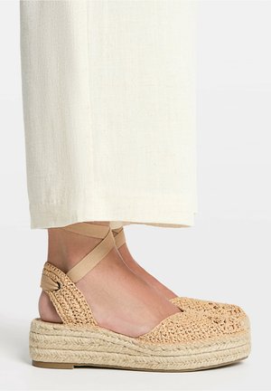 Vævede beige platform espadrille-sko med ankelbånd båret med lyse cremefarvede bukser med vide ben på fødder mod en ensfarvet baggrund.