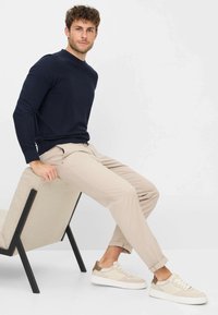 Marine crew-neck sweater, beige slim-fit broek en beige sneakers met bruine accenten. Model zit op een lichtgekleurde stoffen stoel met zwarte poten.
