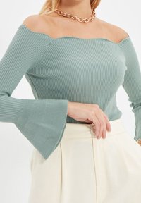 Rippstrick, schulterfreier Pullover in sanftem Mintgrün, mit Glockenärmeln und taillierter Passform. Kombiniert mit cremefarbigen Hosen.
