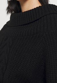 Black knitted sweater with a ribbed turtleneck design. Features a textured pattern, showcasing a cable knit on one side.