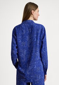 Γυναίκα με μακριά καστανά μαλλιά, φορώντας μπλε πουκάμισο με μακριά μανίκια και παντελόνι με μοτίβο πεϊσλί, γυρισμένη πλάτη σε λευκό φόντο.