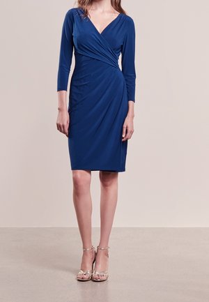 Kvinde iført en knælængde, langærmet, dybblå slå-om-kjole med rynkede detaljer, kombineret med metalliske åbne, høje hæle sandal.