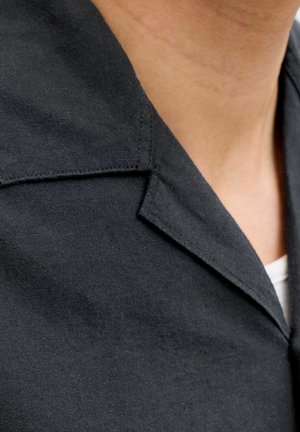 Collo e area superiore del petto che mostrano un blazer in tessuto nero indossato sopra una camicia bianca su una persona.