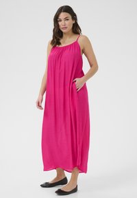 Robe maxi en rose vif, en tissu léger. Présente des bretelles fines, un corsage froncé et des poches latérales. Chaussures de ballet noires plates.