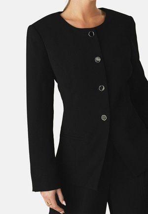 Kvinde iført en tætsiddende sort blazer med fire sølvfarvede sekskantede knapper og lange ærmer, mod en enkel lys baggrund.