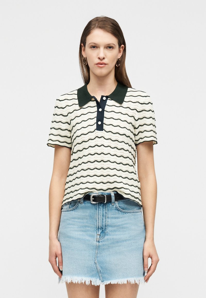 Γυναίκα με ίσια καστανά μαλλιά φορώντας ένα λευκό και μαύρο ριγέ πουκάμισο με κοντά μανίκια και γιακά και φούστα από ανοιχτό τζιν με ξεφτισμένες άκρες.