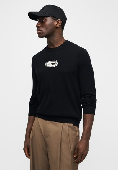 Black knit sweater with a round neck and ribbed cuffs, featuring a white embroidered "VOLTAIRE" logo on the chest. Brown pleated trousers.