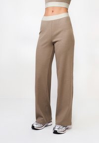 Beżowe spodnie o szerokich nogawkach wykonane z teksturowanej tkaniny, z lekkim pasem w talii z logo "LAMANIA". Połączone z srebrnymi butami do biegania.