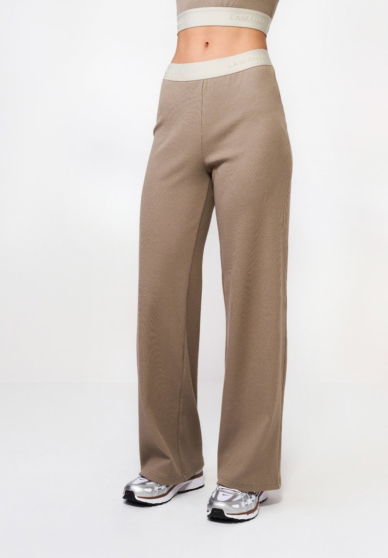 Beżowe spodnie o szerokich nogawkach wykonane z teksturowanej tkaniny, z lekkim pasem w talii z logo "LAMANIA". Połączone z srebrnymi butami do biegania.