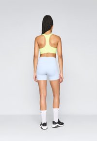 Αθλητικό σουτιέν σε νεόν κίτρινο χρώμα και ανοιχτό μπλε σορτς ψηλής μέσης. Απαλή, ελαστική ύφανση. Το μοντέλο φοράει μαύρα αθλητικά παπούτσια και άσπρες κάλτσες.