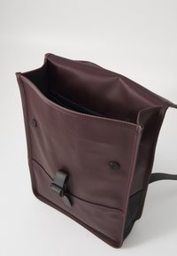 Bolso marrón con parte superior abierta, revelando un interior negro. Presenta un cierre de presión y una correa decorativa en la parte delantera.