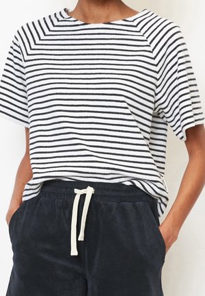 Personne portant une chemise à manches courtes rayée horizontale noire et blanche, rentrée dans un pantalon noir à cordon, les mains dans les poches.