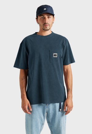 Homme portant une casquette bleu foncé, un t-shirt bleu foncé avec un logo sur la poche avant, et un jean bleu clair, debout devant un fond uni.