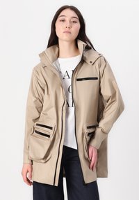 Beżowa wodoodporna kurtka z wysokim kołnierzem, wieloma zamykanymi na zamek kieszeniami i długimi rękawami. Charakteryzuje się luźnym krojem i gładką fakturą.