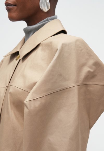 Beige oversize bunda s límečkem, vyrobená z hladké tkaniny. Šedý rolák pod ní a velký stříbrný náušník jako doplněk.