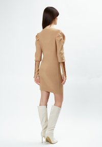 Beige gebreide jurk met pofmouwen, een getailleerde pasvorm en details op de rug, gecombineerd met hoogglanzende witte laarzen tot de knie.