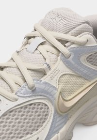 Κοντινό πλάνο ενός μπεζ και ανοιχτογκρι αθλητικού παπουτσιού Nike με πλέγμα, μαξιλαρωμένη γλώσσα και εκρού κορδόνια δεμένα σε σταυρωτό σχέδιο.