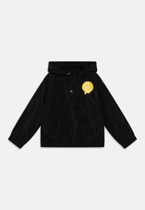 Chaqueta cortavientos negra con capucha, que presenta un gráfico de cara sonriente amarilla en el pecho, puños elásticos y un acabado ligero y brillante.