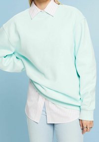 Sudadera de gran tamaño en verde menta con puños acanalados, superpuesta sobre una camisa de cuello a rayas en rosa claro y blanco, combinada con jeans de color azul claro.