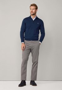 Suéter de cremallera azul marino hecho de tela suave, pantalones grises y zapatos oscuros. Los detalles incluyen un cuello de contraste y un logo bordado en el pecho.