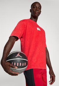 Mand iført en rød Jordan t-shirt og shorts holder en sort og grå Jordan basketball mod en ensfarvet baggrund.
