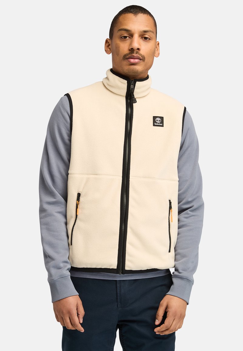 Gilet in pile beige con bordi neri e tirazip arancioni; presenta un colletto alto e tasche con zip. Finitura in tessuto testurizzato.