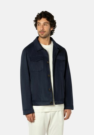 Man met krullend haar, gekleed in een marineblauw jasje met knopen over een wit overhemd en witte broek, kijkt opzij en staat tegen een effen achtergrond.