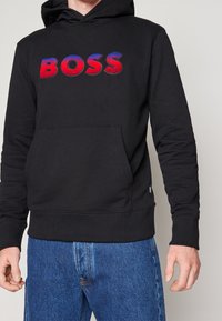 Felpa nera con cappuccio e tasca a marsupio. Presenta il logo "BOSS" in rosso e blu sul petto. Tessuto morbido, polsini e orlo a costine.