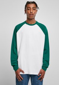Langarm-Raglan-T-Shirt mit einem weißen Körper und grünen Ärmeln. Hergestellt aus Baumwolle, hat es einen Rundhalsausschnitt und eine lockere Passform. Auffällige Nahtdetails.