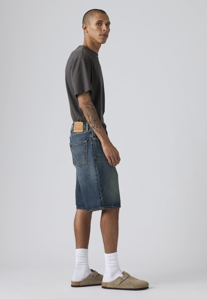 Homme avec tatouage sur le bras portant un t-shirt gris foncé, un short en jean jusqu'aux genoux, des chaussettes blanches et des chaussures beiges à enfiler, debout de profil sur un fond gris.