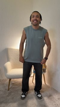 Mand med kort flettehår iført ærmeløs grå skjorte, sorte bukser, sneakers og smartur, stående i et oplyst rum med stol og lille bord.