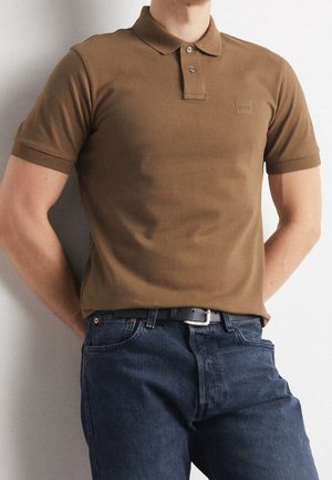 Man draagt een bruine polo met korte mouwen, ingestopt in een donkerblauwe spijkerbroek met een zwarte riem, staand tegen een lichte muur.