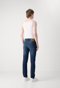 Jeans de mezclilla azul ajustados con un parche de cuero marrón y costuras en contraste, combinados con una camiseta sin mangas blanca ajustada y zapatillas blancas.