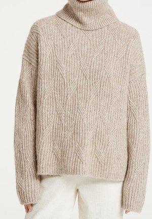 Beige chunky gebreide coltrui met kabelpatroon, lange mouwen en een ontspannen pasvorm, gedragen met witte geribbelde broek.