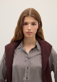 Chemise à rayures marron et blanches avec boutons, associée à un gilet en maille bordeaux posé sur les épaules ; texture lisse, coupe régulière.