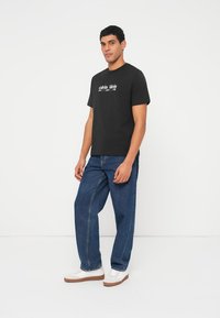 Juoda medvilninė marškinėliai su baltu atspausdintu logotipu, derinami su mėlynais džinsais ir baltais sportbačiais su ruda padu.