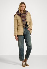Jachetă pufosă bej cu model cusut vertical, eșarfă multicolorată cu dungi, bluză închisă, blugi lejeri albaștri și teniși maro deschis.