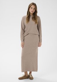 Mujer vestida con un suéter beige de manga larga y una falda a la pantorrilla a juego, con zapatillas marrones, de pie frente a un fondo blanco liso.