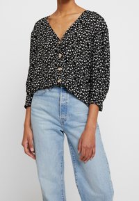 Blusa nera con bottoni e motivo floreale bianco, scollatura a V e maniche 3/4. Abbinata a jeans azzurri a vita alta.