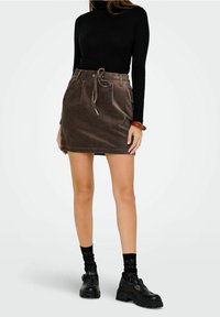 Minigonna marrone in velluto a coste con vita annodata, abbinata a una maglia nera a maniche lunghe e scarpe nere robuste. Dettaglio del tessuto testurizzato visibile.