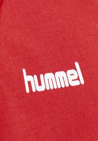 Telas rojas con una textura suave que presentan el logo de la marca "hummel" en blanco, impreso de manera destacada con una tipografía moderna y en negrita.