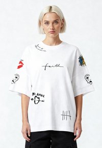 Balta per didelė marškinė su įvairiais atspaudais, įskaitant lūpas, veidus ir tekstą. Su trumpomis rankovėmis ir laisvu siluetu.