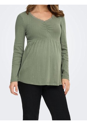 Pregnant woman wearing a ribbed olive green long-sleeve maternity top and black pants standing against a white background.