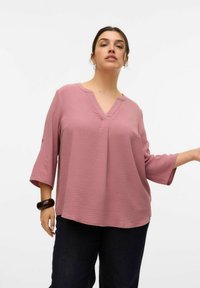 Top rosa loose-fitting e texturizzato con scollo a V e maniche a tre quarti, abbinato a jeans scuri e a un bracciale voluminoso.