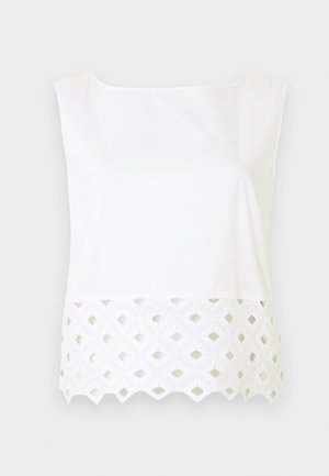 Witte mouwloze top met boothals en kanten zoom met diamantvormige uitsnijdingen op een lichtgrijze achtergrond.