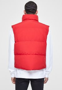 Rote Puffweste mit hohem Kragen, ausgestattet mit horizontalen, gesteppten Abschnitten. Über einem weißen Sweatshirt getragen, mit glatter, matter Textur.